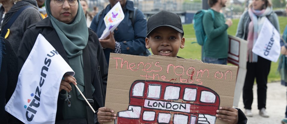 A young boy grins and holds a sign with a red bus, a woman in hijab next to him holds a citizens uk flag