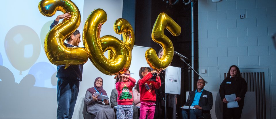Three children are holding golden balloons representing numbers 2635 on stage at a South London Listens Assembly.