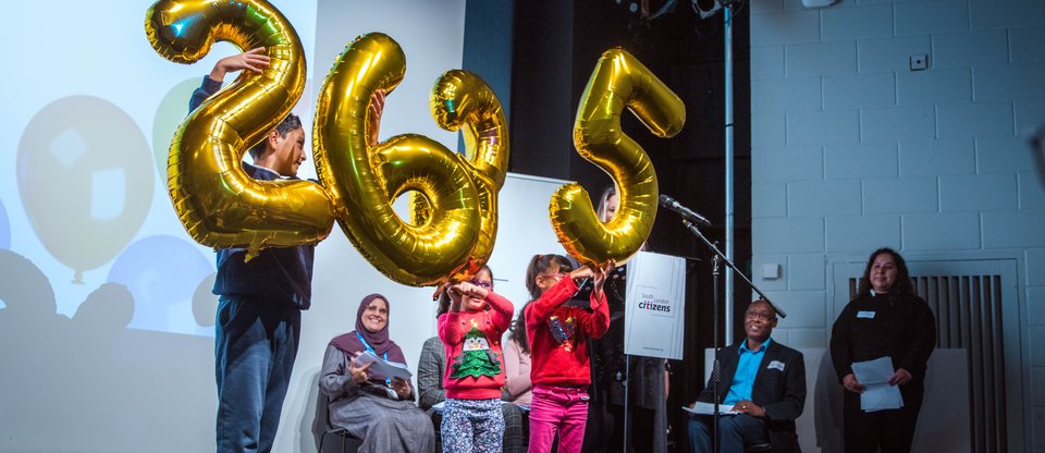 Young children laughing and holding up gold balloons on stage which spell out the number 2635, referring to new living wage employers.