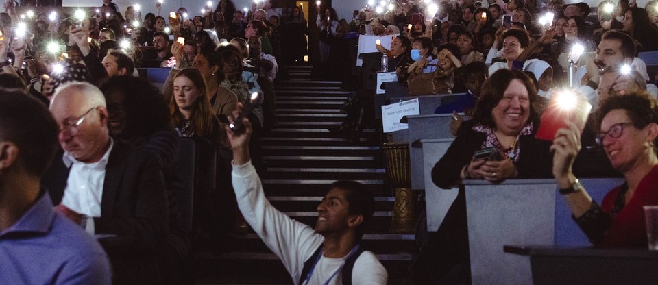 People sit in a dark hall holding up their phone torches smiling at looking towards the stage. A man sits in the middle of the aisle on the stairs.