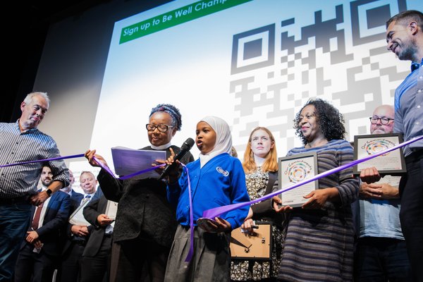 Young leader talks on stage at a South London Listens assembly. She holds a purple ribbon connecting with people from different organisations across the stage.