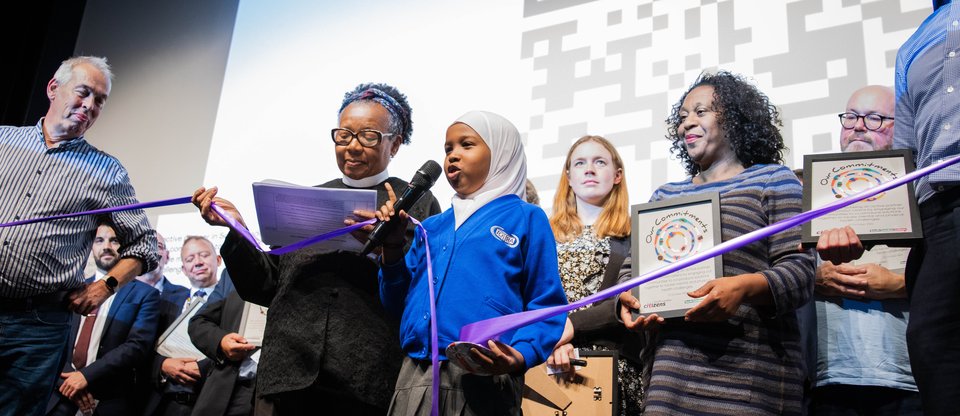 Young leader talks on stage at a South London Listens assembly. She holds a purple ribbon connecting with people from different organisations across the stage.