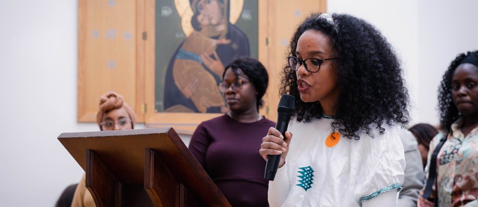 Woman stands on stage in front of parent community leaders and speaks into microphone.