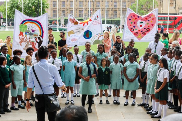 Around 40 primary school children from St Antony's Catholic Primary school stand singing outside in Parliament Square in a semi circle. In front of them is a teacher conductor and a crowd watching. Some of the children are holding up big love heart signs.