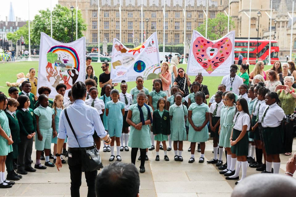 Around 40 primary school children from St Antony's Catholic Primary school stand singing outside in Parliament Square in a semi circle. In front of them is a teacher conductor and a crowd watching. Some of the children are holding up big love heart signs.