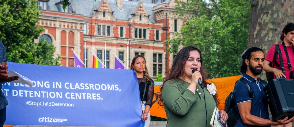 A leader speaking at the child detention action at Parliament Square. They stand with a microphone in front of a blue banner that says 'Children belong in classrooms, not detention centres'.