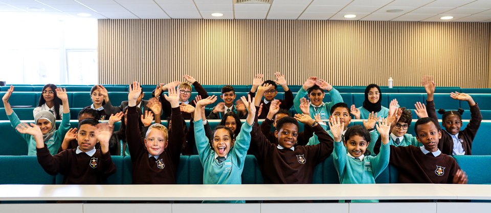 Pupils are sat in two rows looking excited with their hands thrown into the air