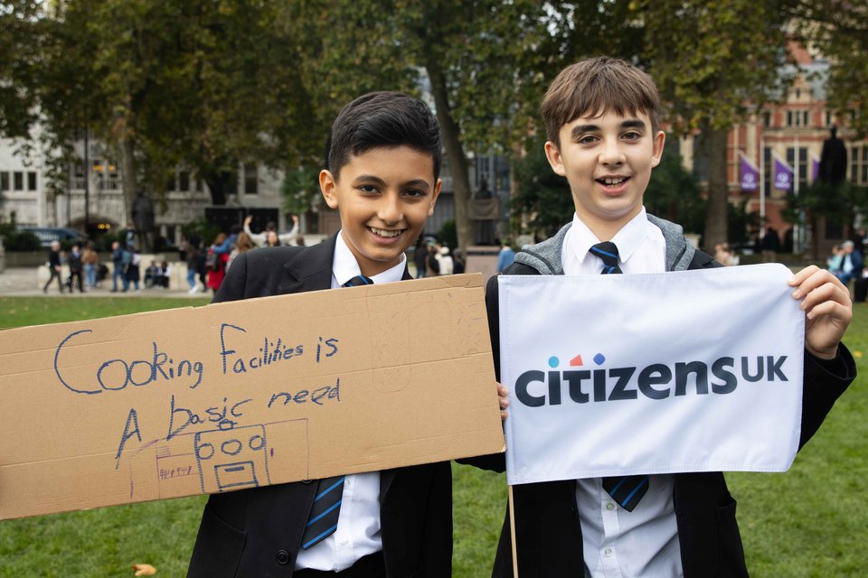 Two young leaders stand in Parliament Square at a housing action, one with a sign that reads "cooking facilities are a basic need" and one with a Citizens UK flag