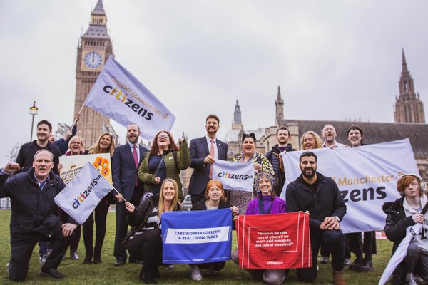 A group of smiling members of Greater Manchester Citizens alliance outside the Houses of Parliament, holding banners to support a real living wage for care workers