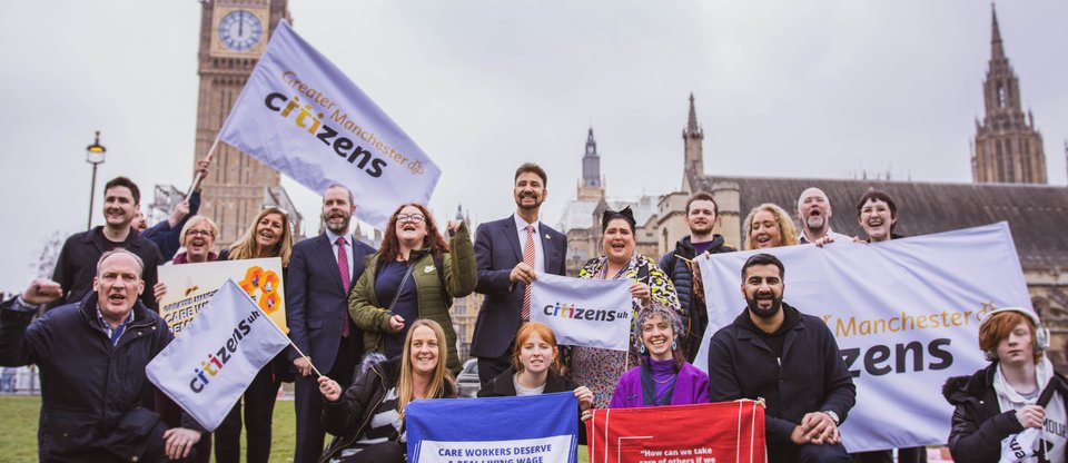 A group of smiling members of Greater Manchester Citizens alliance outside the Houses of Parliament, holding banners to support a real living wage for care workers