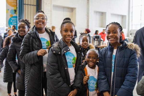 Children wearing Living Wage Foundation t-shirts smile and pose as they queue up to enter a bright warehouse event space, being used for London Living Wage Week's launch event
