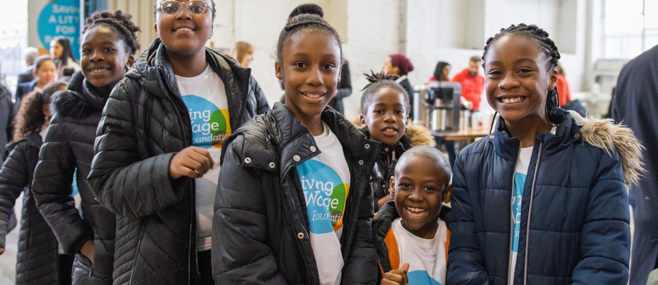 Children wearing Living Wage Foundation t-shirts smile and pose as they queue up to enter a bright warehouse event space, being used for London Living Wage Week's launch event
