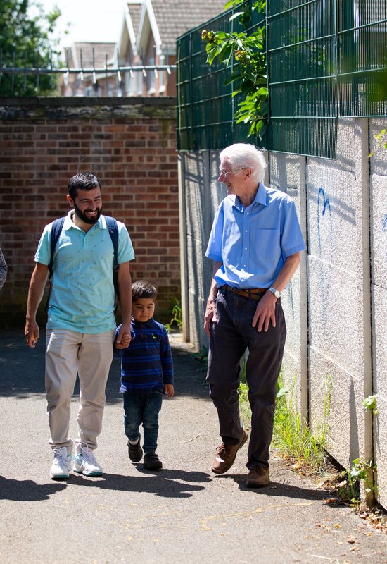 A couple with a young child, holding his father's hand, walks through a bright alleyway. They are smiling and chatting with a gentleman from a Community Sponsorship group.