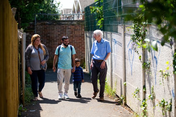 A couple with a young child, holding his father's hand, walks through a bright alleyway. They are smiling and chatting with a gentleman from a Community Sponsorship group.