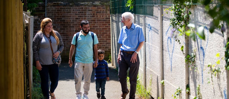 A couple with a young child, holding his father's hand, walks through a bright alleyway. They are smiling and chatting with a gentleman from a Community Sponsorship group.
