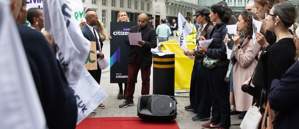 A man with a microphone speaks at an action, with people standing either side of him with flags and placards and the BBC building in the back