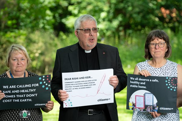 Three community leaders hold signs asking for safer homes. Rev Ian Rutherford remembers those who lost their lives to unfit homes.