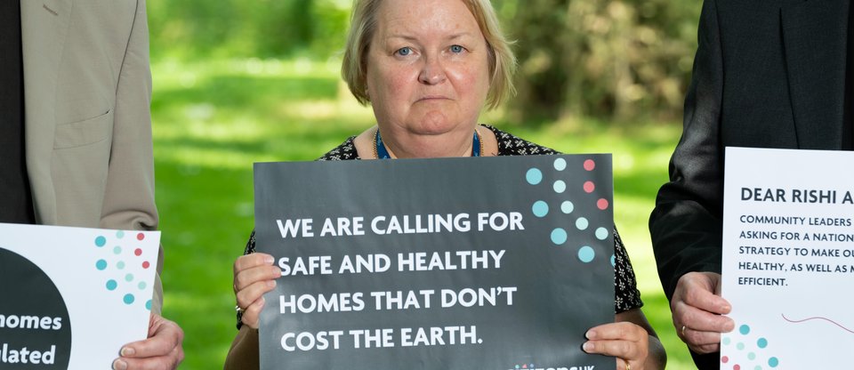 A community leader is holding a sign that reads 'We are calling for safe and healthy homes that don't cost the earth.'