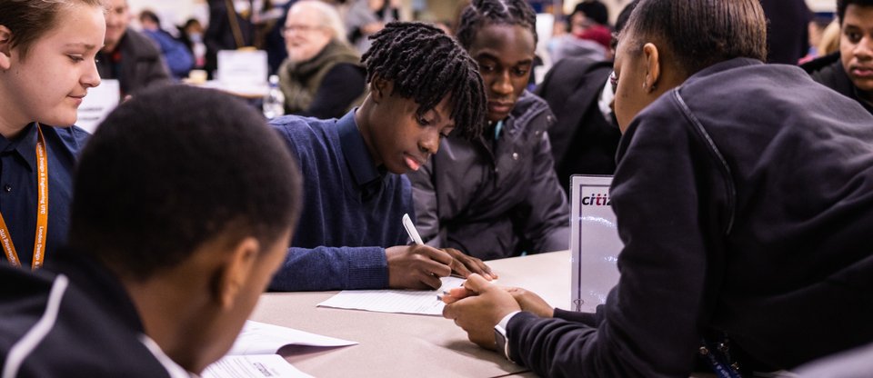 Student writing on piece of paper at a table with other students and an adult