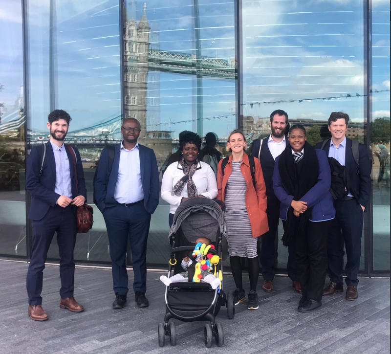 Parent Action leader Elsie and her baby join campaigners from Southwark Citizens on a sunny day outside City Hall. They pose in front of a glassy building with Tower Bridge reflected above them.