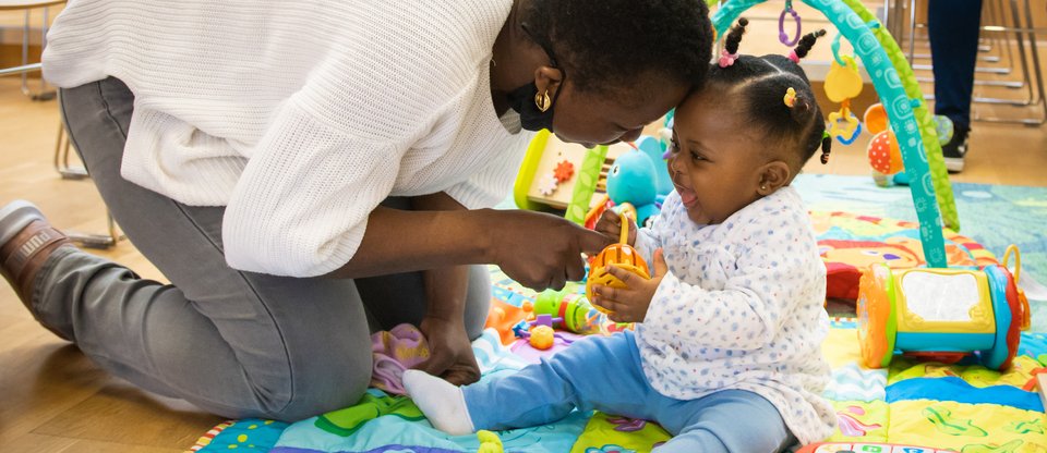 Mum and baby playing together with toys on a blanket at a Parent Action session. The baby is smiling and giggling whilst mum is kneeling down and resting her forehead affectionately against the baby's forehead.