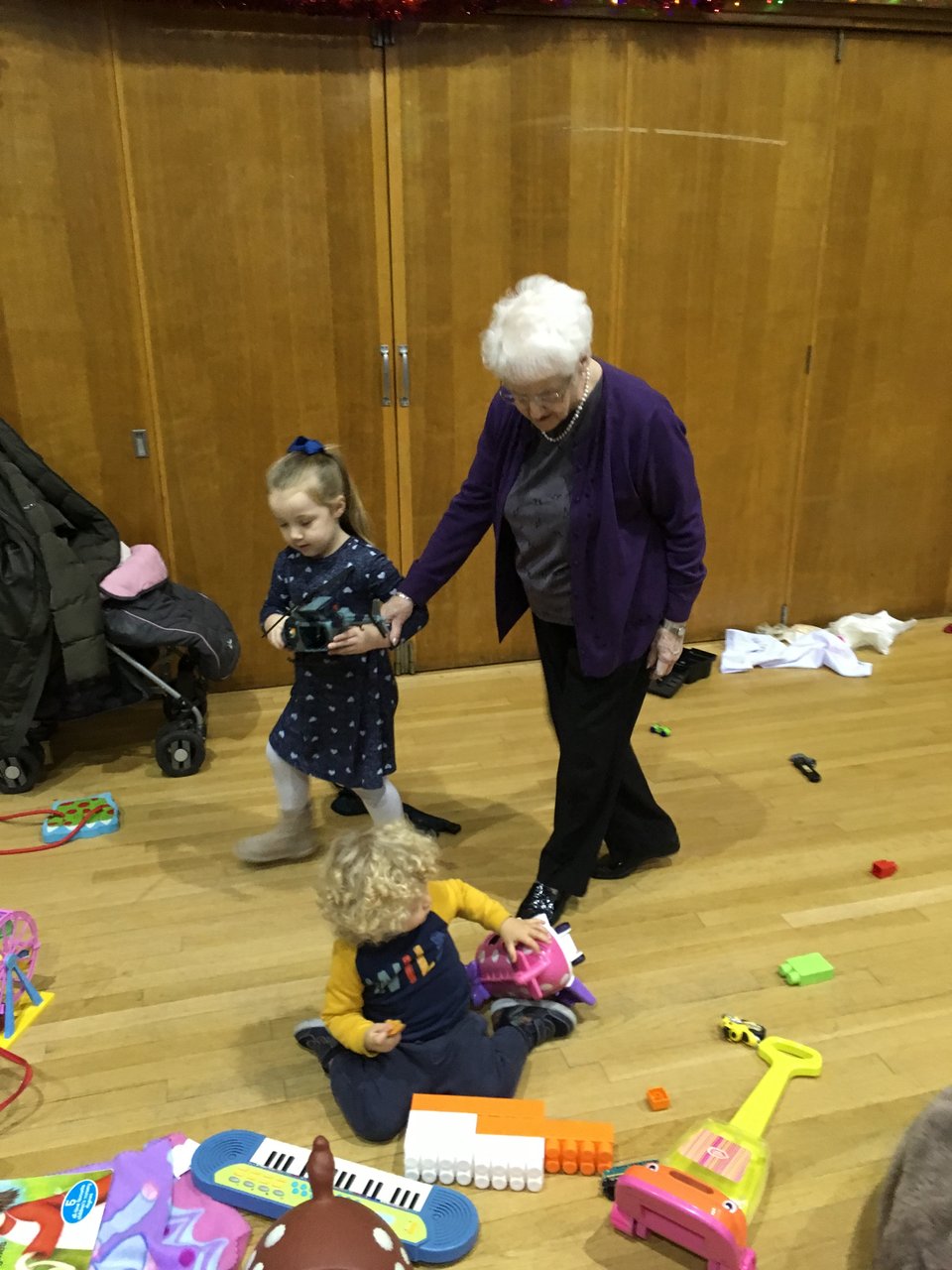 Grandma holds a child's hand as they walk around a room filled with toys