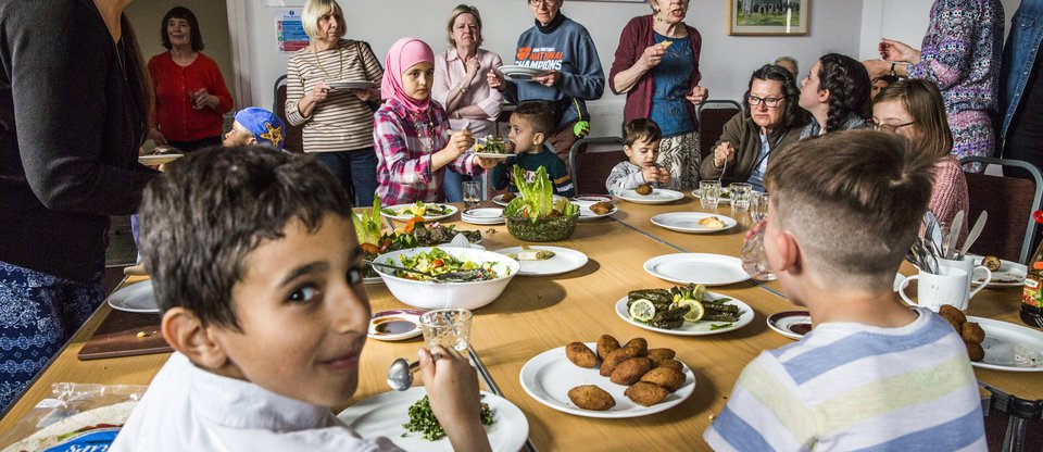 Children and adults eating together around a large table with a small boy smiling at the camera