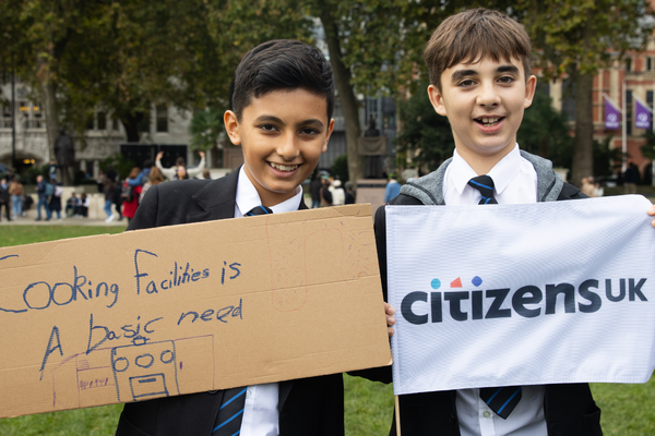 Two young pupils holding signs up, on the left is 'cooking facilities is a basic need' and on the right is a Citizens UK flag