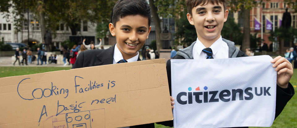 Two young pupils holding signs up, on the left is 'cooking facilities is a basic need' and on the right is a Citizens UK flag