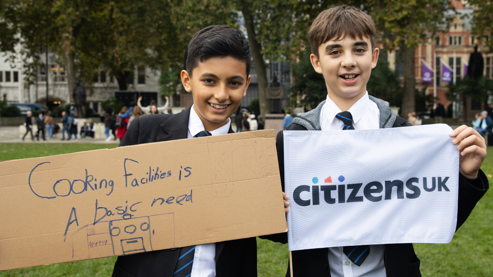 Two young pupils holding signs up, on the left is 'cooking facilities is a basic need' and on the right is a Citizens UK flag