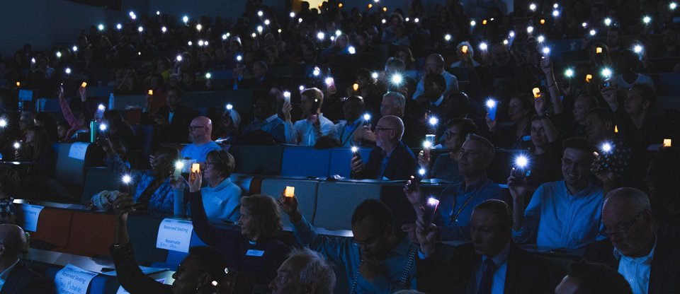 A room full of leaders hold their phone lights up in a semi-darkness at an assembly