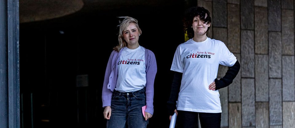 Two young female leaders with Tyne and Wear Citizens t-shirts stand outside facing the camera. One confidently places her hands on hips.