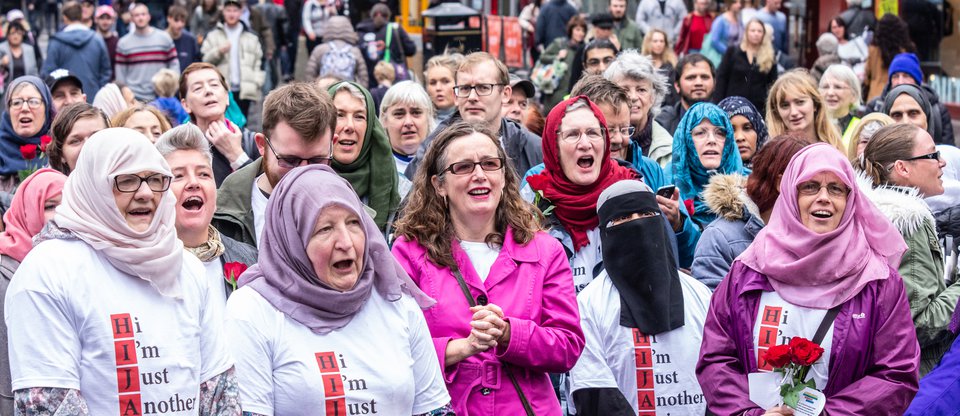 Group of people singing together with T-shirts saying "Hi I'm Just Another Being" spelling out hijab