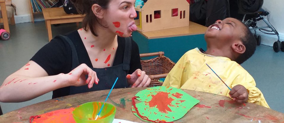 Parent community leader with paint on her face sits at an arts and crafts table with a young child. The child is holding a paint brush and leaning back in hysterics with a big grin on their face, as the woman sticks out her tongue.
