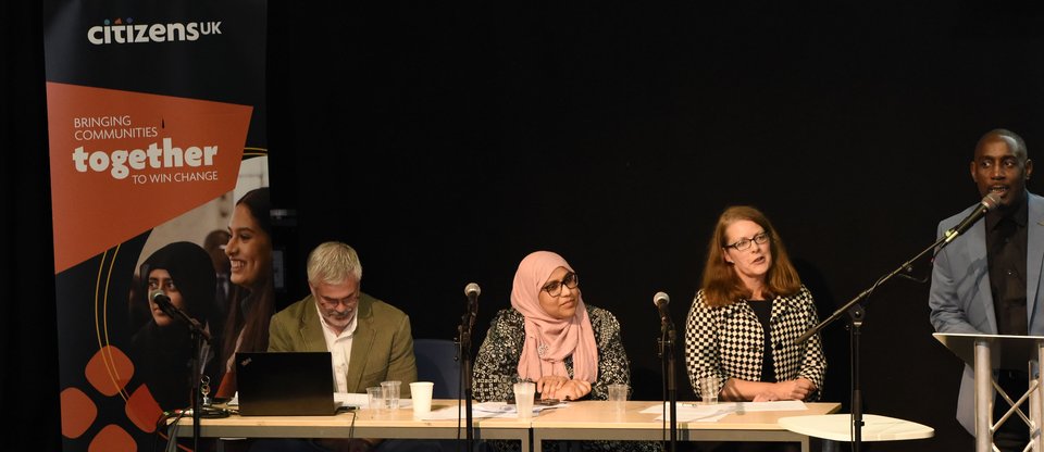 Three community leaders sit at a panel whilst the fourth speaks on stage behind a microphone, at a Birmingham Citizens event.