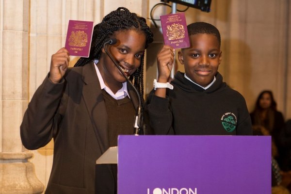Two children stand behind a London Citizens pillar at an event, holding their passports in the air and smiling