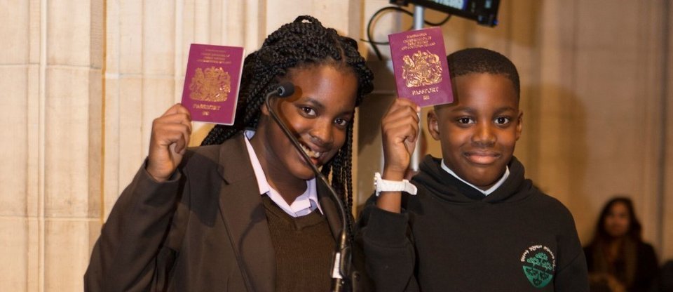 Two children stand behind a London Citizens pillar at an event, holding their passports in the air and smiling