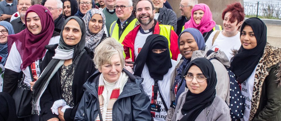 A multi-faith group of people standing together by the sea holding roses and wearing t-shirts taking action against hate crime