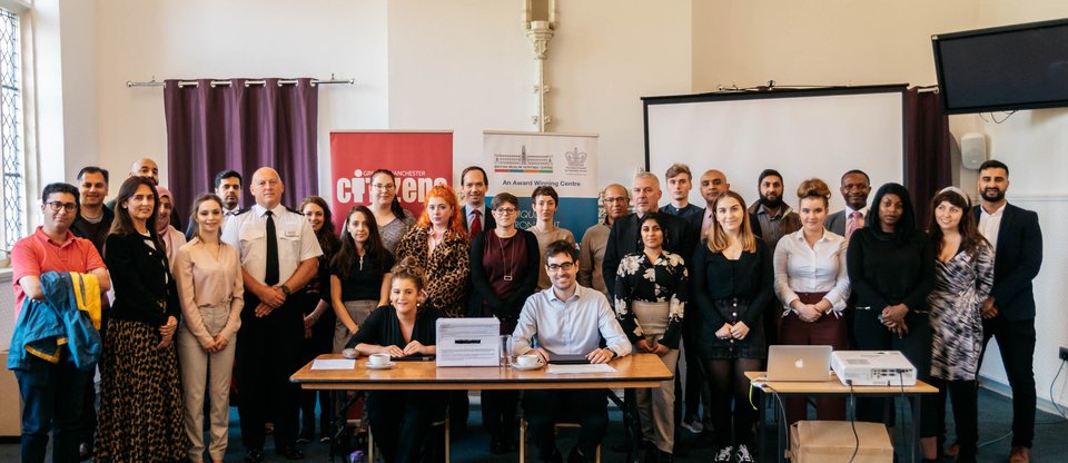 Community leaders, including hate crime survivors, faith groups, women's organisations, university students, pupils, teachers and more stand around a table in Greater Manchester. Seated at the table are representatives from the Law Commission.