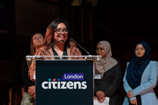 A Citizens UK leader is smiling and speaking behind a lectern with the London Citizens logo.