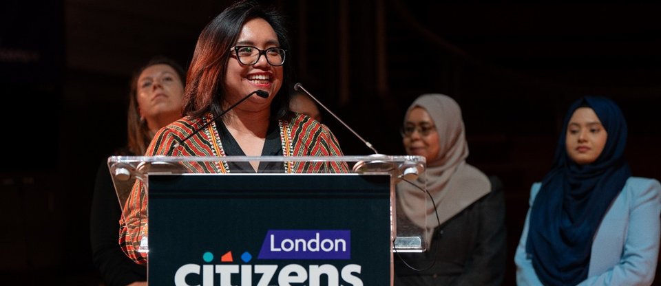 A Citizens UK leader is smiling and speaking behind a lectern with the London Citizens logo.