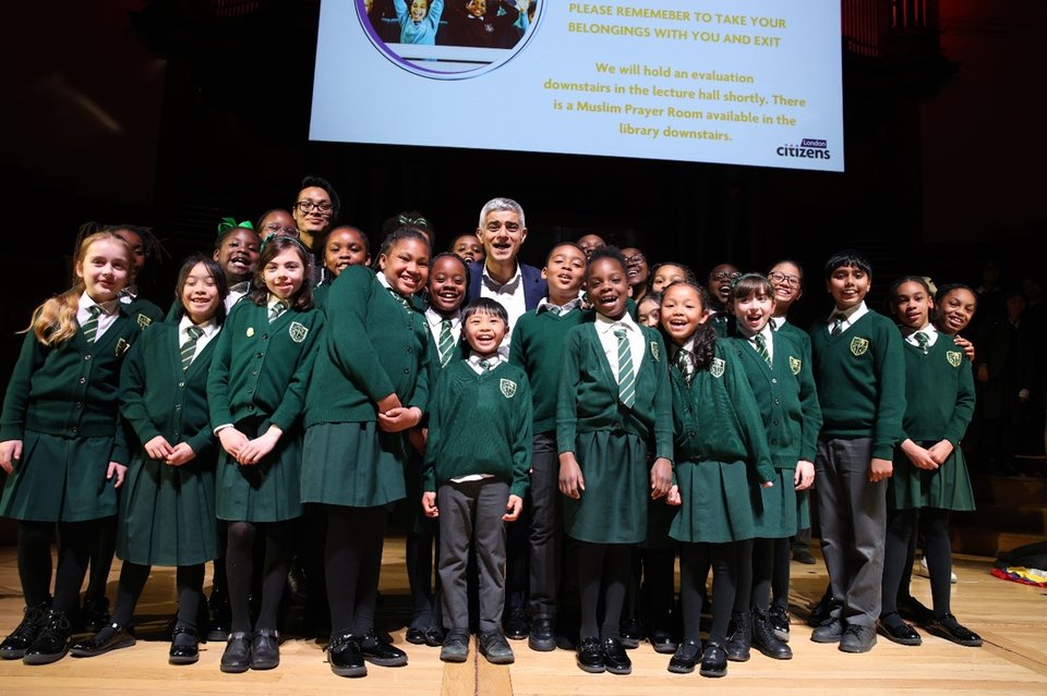 Sadiq Khan is standing on stage with a choir of young people at a London Citizens Assembly.