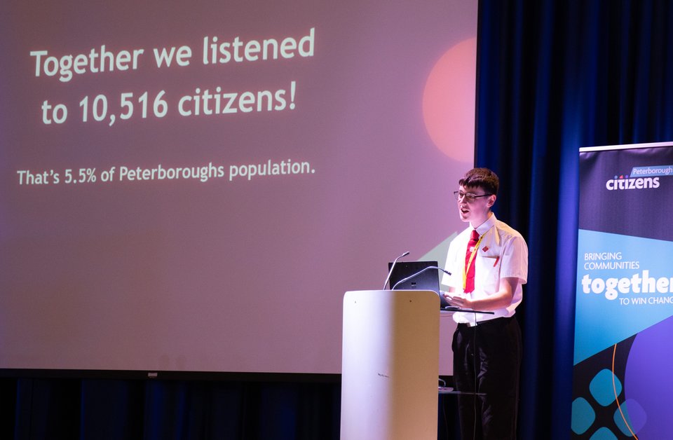 Young school boy stands on stage at a podium, looking out to the audience with a presentation slide behind him reading "together we listened to 10,516 citizens! That's 5.5% of Peterborough's population".