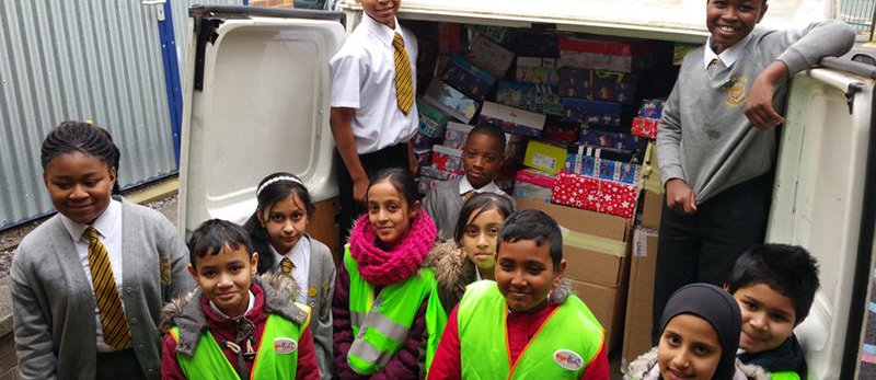 11 school children from a Refugee Welcome school stand smiling around a van filled with presents and gifts