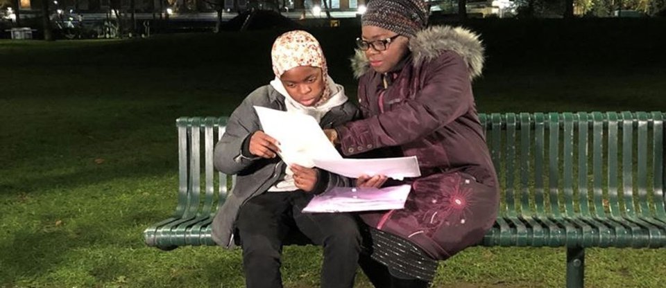 A woman and young girl sit on a park bench at night time, being filmed for a Sky News interview. They are looking at paperwork and are lit up by streetlights.