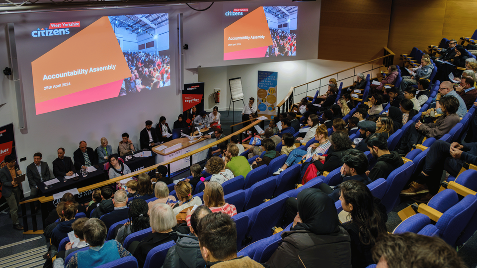 Birds eye view shot of lecture theatre filled with people in their seats, facing the front of the room which has a panel of 12 people sitting across a long table